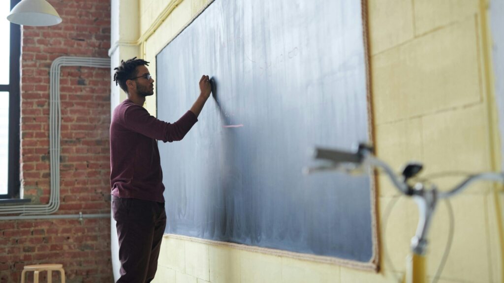 Man with dreadlocks writing on a blackboard in a classroom setting.
