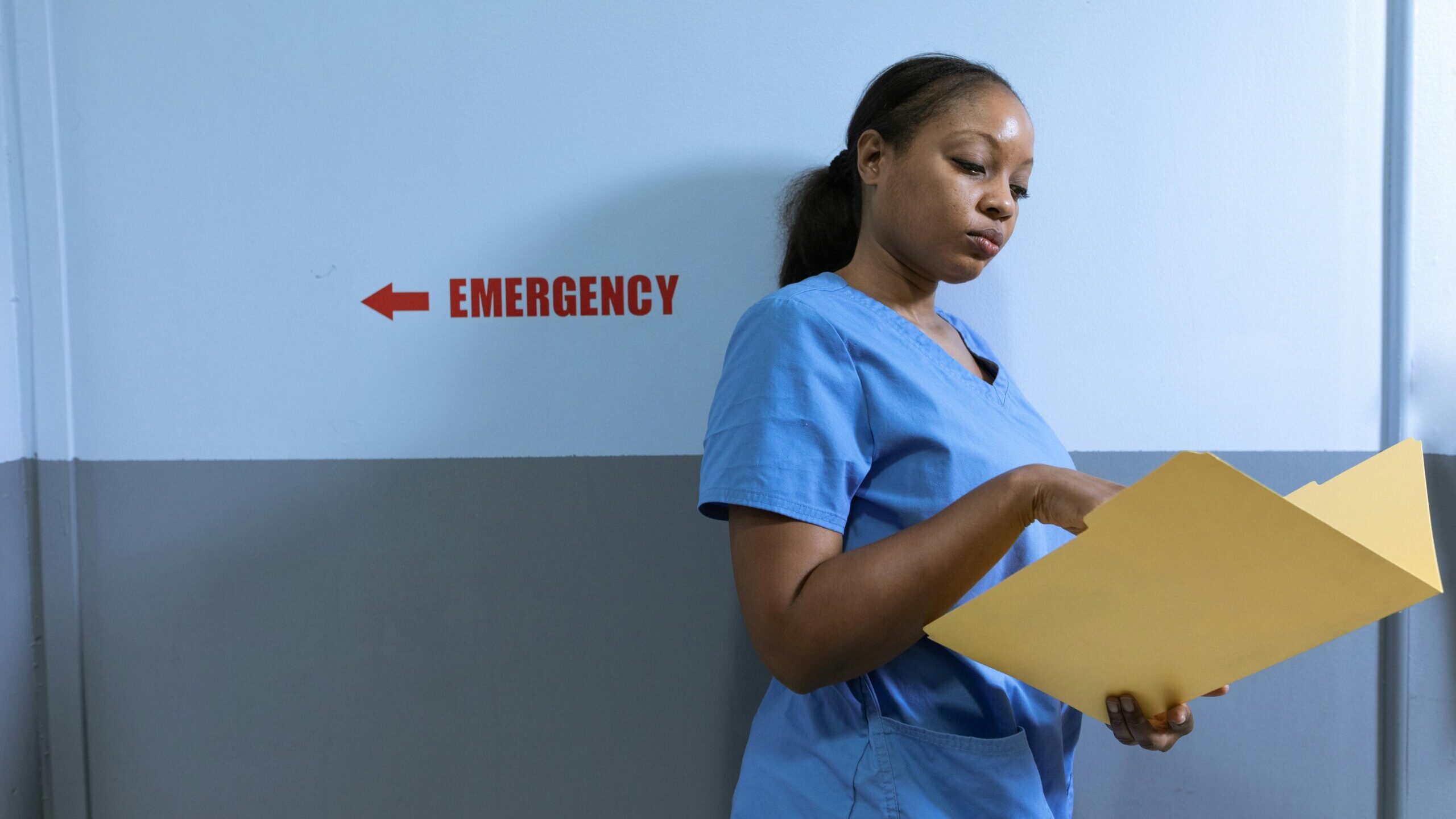 A nurse in blue scrubs reads a folder near an emergency room sign in a hospital.